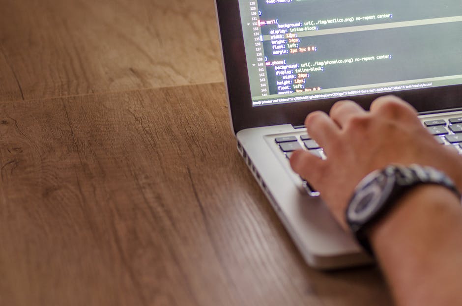This image shows a close-up view of a person working on a laptop placed on a wooden surface. The person's hand, wearing a watch, is positioned over the keyboard, interacting with the screen that displ